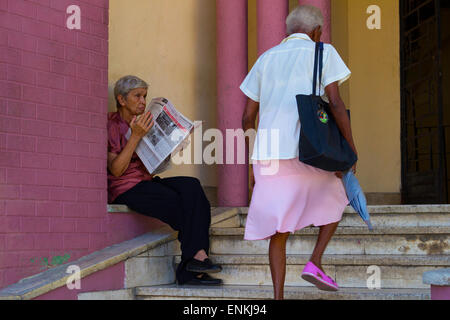 Ältere Frau sitzt vor Haus lesen kommunistischen kubanische Tageszeitung und Frau mit Regenschirm vorbei Stockfoto