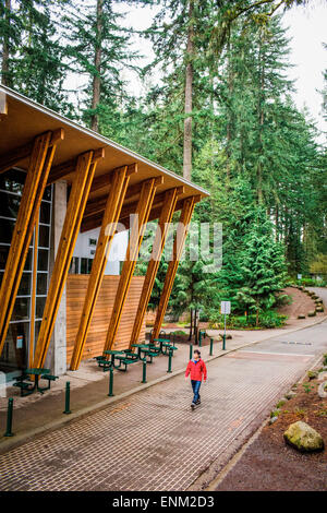 VANCOUVER, BRITISH COLUMBIA, KANADA. Ein Mann in einem roten Mantel und Jeans geht neben einem modernen Besucherzentrum in einem bewaldeten Park. Stockfoto