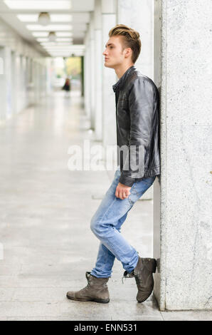 Attraktive blonde junge Mann mit Lederjacke draußen gegen Pfeiler, wegschauen Stockfoto