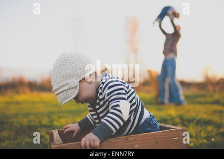 Ein Baby setzt sich im Inneren eine alte Holzkiste während ihr Vater eines ihrer allerhöchsten in die Luft hebt. Stockfoto