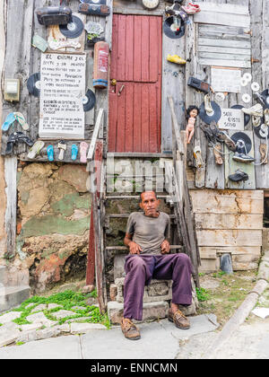 Ein Hispanic kubanischen sitzt Man auf den Stufen von seinem sehr seltsam, eingerichtete unkonventionell Holzhaus. Stockfoto