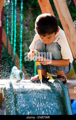 Gemischtes, kaukasisch-asiatisches Kind, Junge, 7-8 Jahre alt, kniend auf der überdachten Rutsche, Sprühen der Rutsche sauber mit einem Schlauch Rohr im Garten. Stockfoto