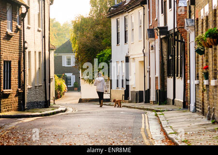 Eine eingefleidete Frau in der Ferne, die während der goldenen Stunde bei Sandwich in Kent vom Betrachter weg läuft, während sie ihren Hund in einer leeren Dorfstraße führt. Stockfoto