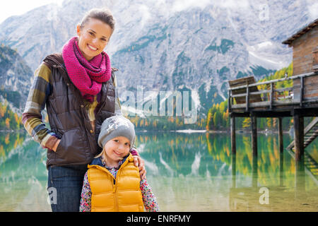 Eine Mutter und Tochter Stand lächelnd an den Ufern des Sees Bries. Hinter ihnen bietet das Stille Wasser perfekter Spiegelreflexionen auf die Berge, Herbstlaub und die hölzerne Pier. Stockfoto