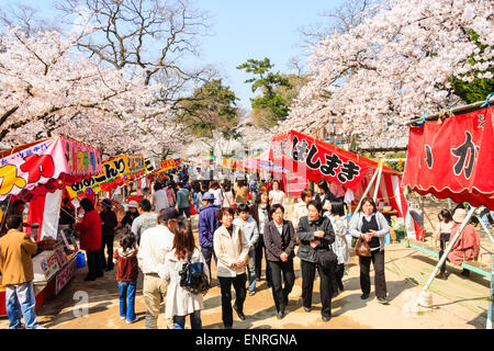 Menschenmassen, die am Flussufer entlang zwischen zwei Reihen von Speise- und Spielständen während des frühlingshaften Kirschblütenfestes in Japan spazieren. Stockfoto