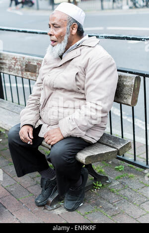Asiatische tragen Topi Kopf saß in Salisbury Street Wear Rauchen einer Zigarette auf der Straße Sitzbank Stockfoto