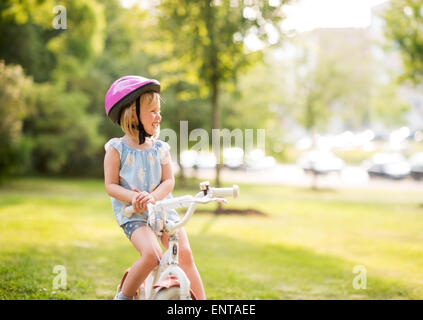 Ein junges Mädchen sitzt auf ihr Fahrrad in einem Stadtpark, lächelnd, Ruhe und glücklich. Einen rosa Helm zu tragen, weiß sie, wie man b üben Stockfoto