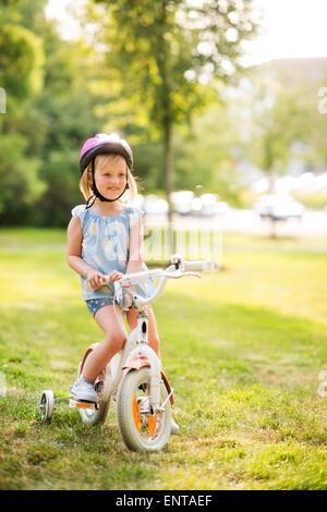 Eine junge Blondine sitzt blauäugige Mädchen auf ihr Fahrrad in einem Stadtpark, lächelnd, Ruhe und glücklich. Sie weiß mit rosa Helm, Stockfoto