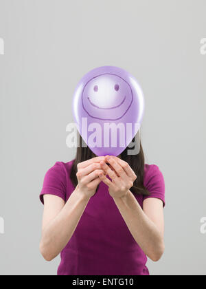 Frau verbirgt ihr Gesicht Behing lila Ballon mit Smiley-Gesicht gezeichnet, auf grauem Hintergrund Stockfoto