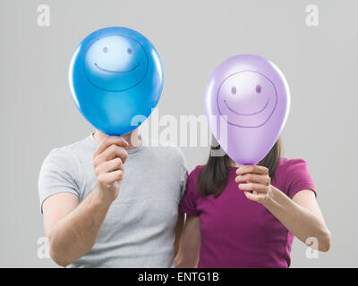 paar, die ihre Köpfe hinter bunten Luftballons mit Smiley-Gesichter vor grauem Hintergrund ausblenden Stockfoto