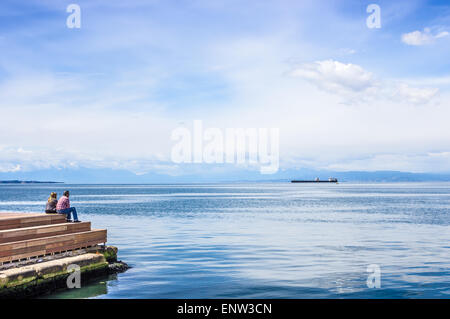 Paar mittleren Alters sitzen auf Pier vor Tanker auf das tiefblaue Meer und mythologischen Mount Olympus am Horizont Stockfoto