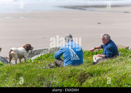 Zwei Männer saßen im Mai mit Hund am Strand von Whitesands Bay, Pembrokeshire Coast National Park, Wales UK Stockfoto