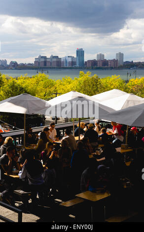 USA, New York, Manhattan, Menschen, die an den Tischen unter Sonnenschirmen an einem Essen Ort am Chelsea Markt Übergang auf die lineare High Line Park auf einem stillgelegten erhöhte Eisenbahn Sporn die Westseite Linie neben dem Hudson River genannt. Stockfoto