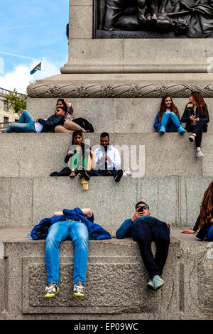 Menschen sitzen auf der Basis des Nelson Säule auf dem Trafalgar Square anlässlich des 70. Jahrestages des VE Tag feiern, London, UK Stockfoto