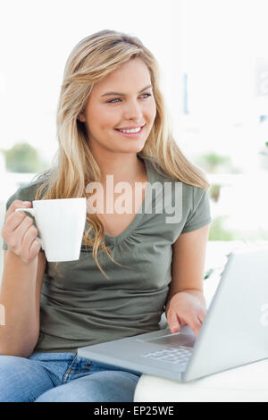 Frau, die leicht zur Seite, als sie eine Tasse in der Hand hält und ihr Laptop nutzt Stockfoto