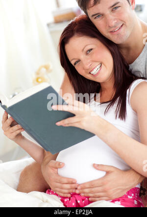 fröhliche zukünftige Eltern Lesen eines Buches sitzt auf ihrem Bett im Laufe des Vormittags Stockfoto