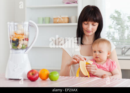 Wunderschöne Brünette Frau eine Banane schälen, halten Sie ihr Baby auf den Knien in der Küche Stockfoto