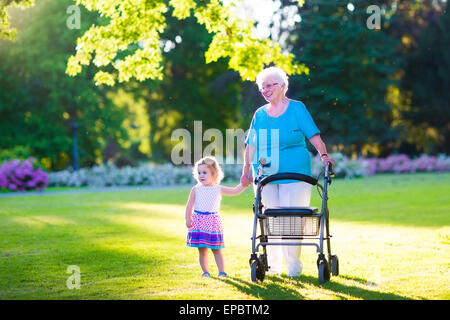 Glücklich senior Lady mit einer Gehhilfe oder Rollstuhl und ein wenig Kleinkind Mädchen, Großmutter und Enkelin, Spaziergang im park Stockfoto