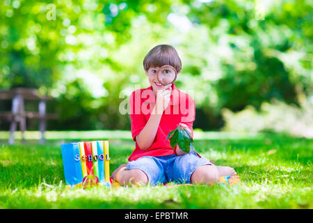 Glücklich Schulkind, Lachen Junge sitzt auf einer grünen Wiese auf dem Schulhof Blick durch eine Lupe, die Blätter zu erkunden Stockfoto