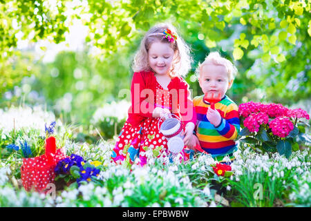 Niedliche lockige kleine Mädchen in einem roten Sommerkleid und lustige Baby junge arbeitet in der Gartenbewässerung ersten Frühlingsblumen Stockfoto