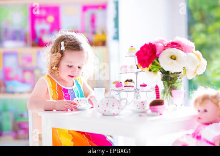 Entzückende Kleinkind Mädchen mit dem lockigen Haar trägt ein buntes Kleid an ihrem Geburtstag spielen Teeparty mit einer Puppe, Spielzeug-Gerichte Stockfoto