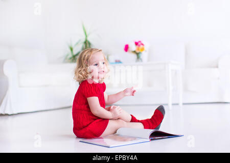 Niedliche kleine Mädchen mit dem lockigen Haar mit einer warmen roten Strickkleid und Socken ein Buch auf dem Boden sitzend Stockfoto