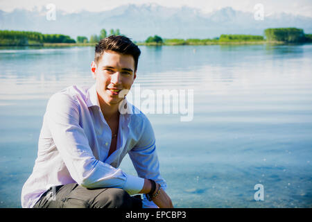 Hübscher junger Mann am Ufer eines Sees an einem sonnigen, ruhigen Tag, sitzen Stockfoto