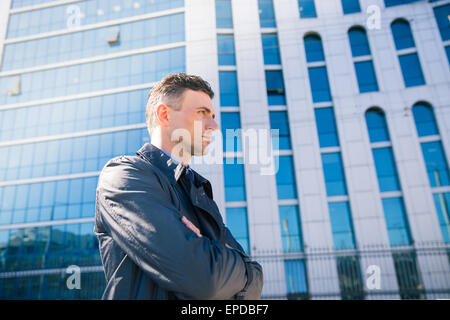 Seite Ansicht Porträt eines Geschäftsmannes stehend mit Armen gefaltet im Freien. Glasbau auf Hintergrund Stockfoto