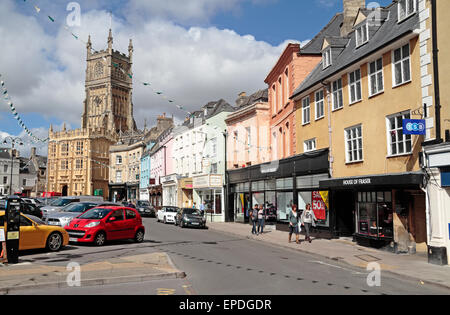 Die Kirche St. Johannes der Täufer und die bunten Gebäude am Marktplatz, Cirencester, Gloucestershire, UK. Stockfoto