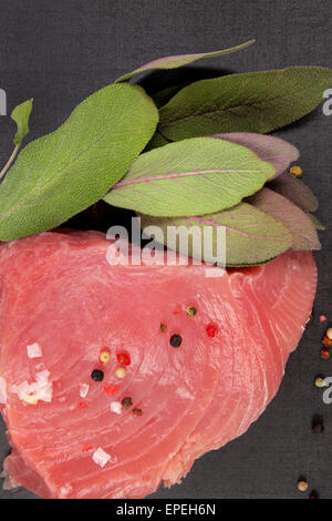 Rohen frischen Thunfischsteak mit frischem Salbei Kräuter, Pfefferkörner und Salz auf schwarzem Hintergrund, Ansicht von oben. Stockfoto