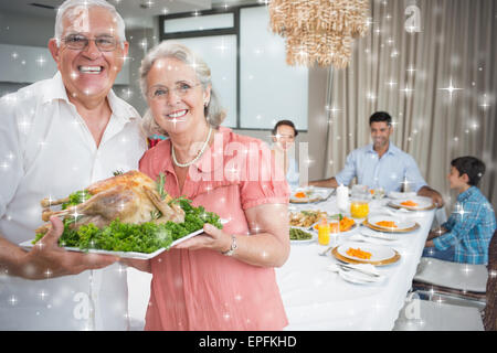 Zusammengesetztes Bild der Großeltern halten Hähnchen braten mit Familie am Esstisch Stockfoto