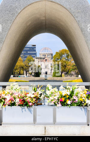 Blick durch das Hiroshima-Kenotaph mit Blumen davor, zur ewigen Friedensflamme und dann zur Atombombenkuppel, Genbaku Domu am sonnendurchfluteten Tag. Stockfoto