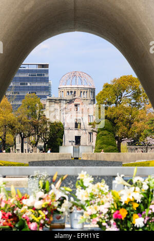 Blick durch das Hiroshima-Kenotaph mit Blumen davor, zur ewigen Friedensflamme und dann zur Atombombenkuppel, Genbaku Domu am sonnendurchfluteten Tag. Stockfoto