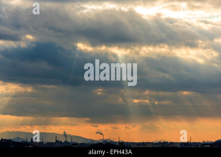 Japan, Himeji. Sunburst durch dicke stürmische dunkle Wolken über der Stadt und dem Industriegebiet mit mehreren hohen rauchenden Fabrikschornsteinen, orangefarbenem Himmel. Stockfoto