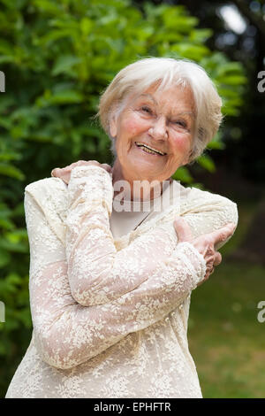 Glücklich lachende Seniorin ziemlich Spitzen oben stehend umarmt sich mit ihren Armen in einem sonnigen Garten. Stockfoto