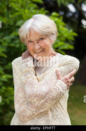 Lächeln freundliche ältere Frau in einem stilvollen Spitzen Bluse halten ihre Arme um sich, während sie in einem sonnigen Sommer-Park steht. Stockfoto