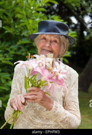 Gerne ältere Dame in einem stilvollen Spitzen Kleid und Hut stehen draußen in ihrem Garten mit einem Bouquet von frischen rosa Lilien. Stockfoto