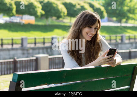 Junge Schönheit Frau viel Spaß mit Smartphone auf der Bank sitzen. Fröhliches kaukasische Mädchen SMS mit Handy im Stadtpark Stockfoto