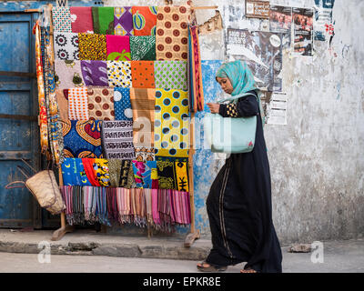 Frau geht neben einem Rack mit bunten Kleidungsstücken namens Kanga und Kitenge in Stonetown, Zanzibar. Stockfoto