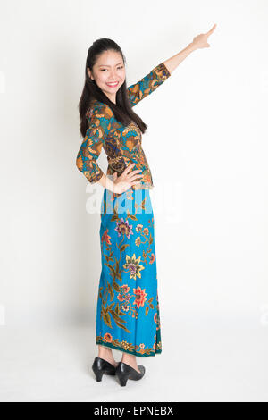 Ganzkörper Portrait glücklich südostasiatische Frau in Batik Kleid Finger Weg zeigen, auf einfarbigen Hintergrund stehend. Stockfoto