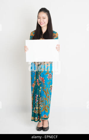 Ganzkörper-Porträt von Southeast Asian Girl in Batik Kleid Hände halten weißen leere Tafel, auf einfarbigen Hintergrund stehend. Stockfoto