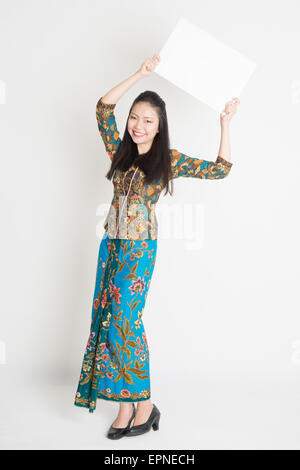 Ganzkörper-Porträt von Southeast Asian Girl in Batik Kleid Hände halten weiße leere Plakat stehen auf einfarbigen Hintergrund. Stockfoto