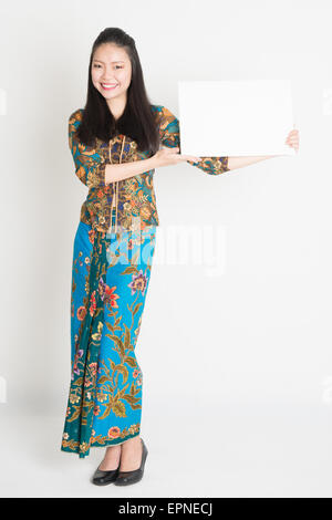 Ganzkörper-Porträt von südostasiatischen Mädchen in Batik Kleid Händen hielt weiße leere Karte, stehend auf einfarbigen Hintergrund. Stockfoto