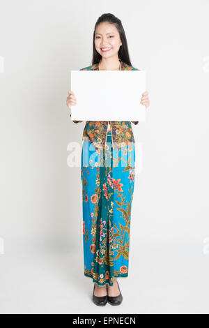 Ganzkörper-Porträt von südostasiatischen Mädchen in Batik Kleid Hände halten weiße leere Plakat, stehend auf einfarbigen Hintergrund. Stockfoto