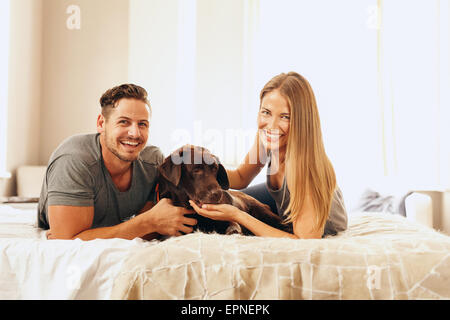 Aufnahme eines jungen Paares auf dem Bett liegend mit ihrem Hund. Fröhlicher Mensch und Frau Blick in die Kamera mit ihrem Haustier im Schlafzimmer. Stockfoto