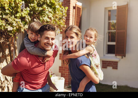 Glücklicher Vater und Mutter Blick in die Kamera, während Sie ihren Sohn und Tochter Huckepack. Glückliche junge Familie von vier Spielen in ihrer Stockfoto