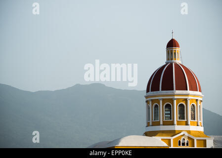 GRANADA, Nicaragua – die Kathedrale von Granada mit ihrem charakteristischen gelb-weißen Äußeren dominiert die Skyline des Parque Central. Die Kuppel der Kathedrale von Granada gegen den Vulkan Mombacho in der Ferne. An dieser Stelle gibt es seit etwa 1525 eine Kirche, die aber in den folgenden Jahrhunderten mehrmals zerstört und wieder aufgebaut wurde, als die Stadt Granada von Piraten und anderen angegriffen wurde. Der Bau der heutigen Version begann 1888, wurde aber erst 1972 vollständig abgeschlossen. Mit seinem markanten Gelb mit weißer Zierleiste, steht es über dem Parque Central im Herzen von GRA Stockfoto