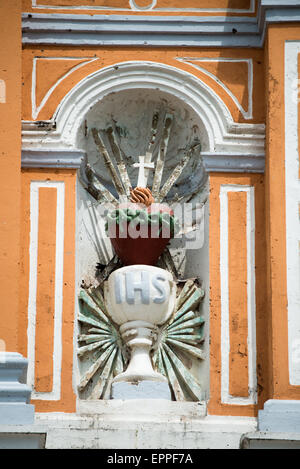 Iglesia De Xalteva Heiligstes Herz IHS Kelch Guszbetonrelief Granada Nicaragua | GRANADA, Nicaragua — Ein bemaltes Relief eines Kelches mit dem IHS-Christogramm, gekrönt von einem flammenden, von Dornen umgebenen Heiligsten Herzen, füllt eine Außenalkoven an der Iglesia de Xalteva. Der Bildhauer Jorge Navas Cordonero ausführte die Außenreliefs der Kirche in einer Mischung aus Guszbeton und Kalk, die poliert wurde, um Marmor zu ähneln, und vollendete das Dekorationsprogramm im frühen zwanzigsten Jahrhundert. IHS ist die Abkürzung für das lateinische Iesus Hominum Salvator, „Jesus, Erlöser der Menschheit.“ Stockfoto