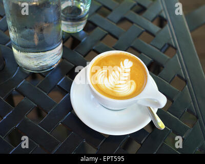 Nahaufnahme von Cappuccino mit Latte Art weiße Tasse und Untertasse mit Flasche und Glas auf Terrassentisch, Kanada Stockfoto