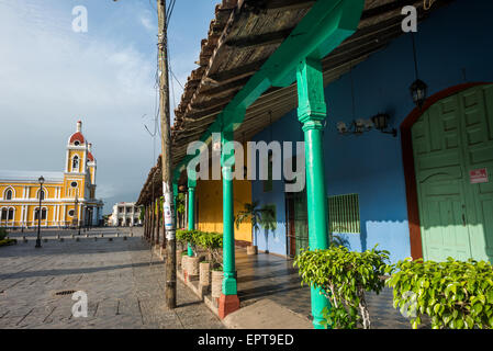 Granada-Kathedrale Bunte spanische Kolonialarchitektur Granada Nicaragua | GRANADA, Nicaragua — Hellgrüne Holzsäulen stützen einen tiefen Kolonialportikus neben dem Parque Central, wobei die gelb-weiße Kathedrale von Granada auf der Plaza sichtbar ist. Das überhängende Terrazzodach und die überdachte Passage spiegeln eine Standardanpassung der spanischen Kolonialarchitektur wider, um Fußgänger vor tropischer Sonne und Regen zu schützen. Der neoklassizistische Umbau der Kathedrale, der 1888 begann, wurde 1972 abgeschlossen. Stockfoto